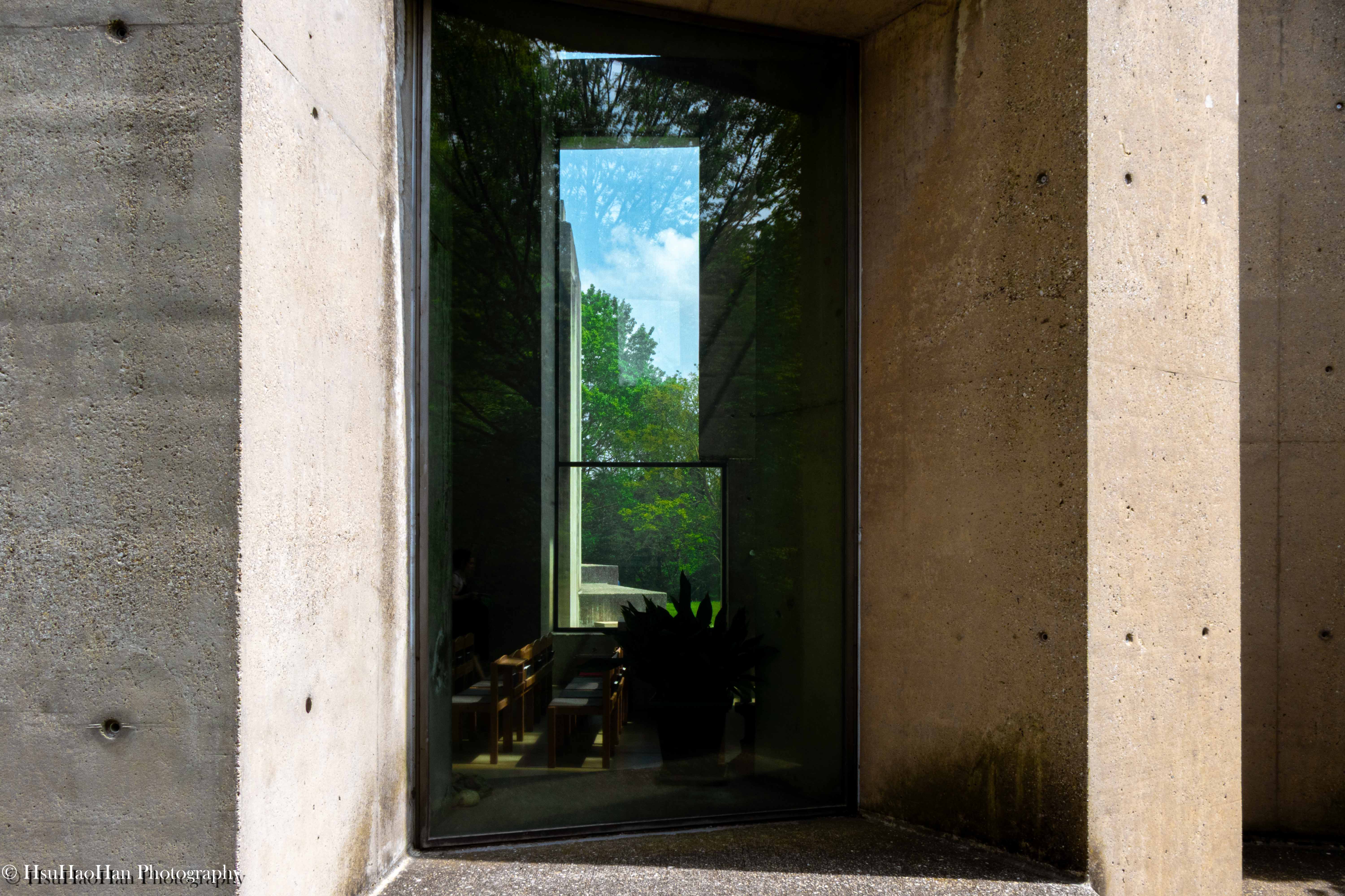 從維也納沃特魯巴教堂的混凝土牆邊，透過玻璃窗看見室內的座椅與室外的綠樹倒影。