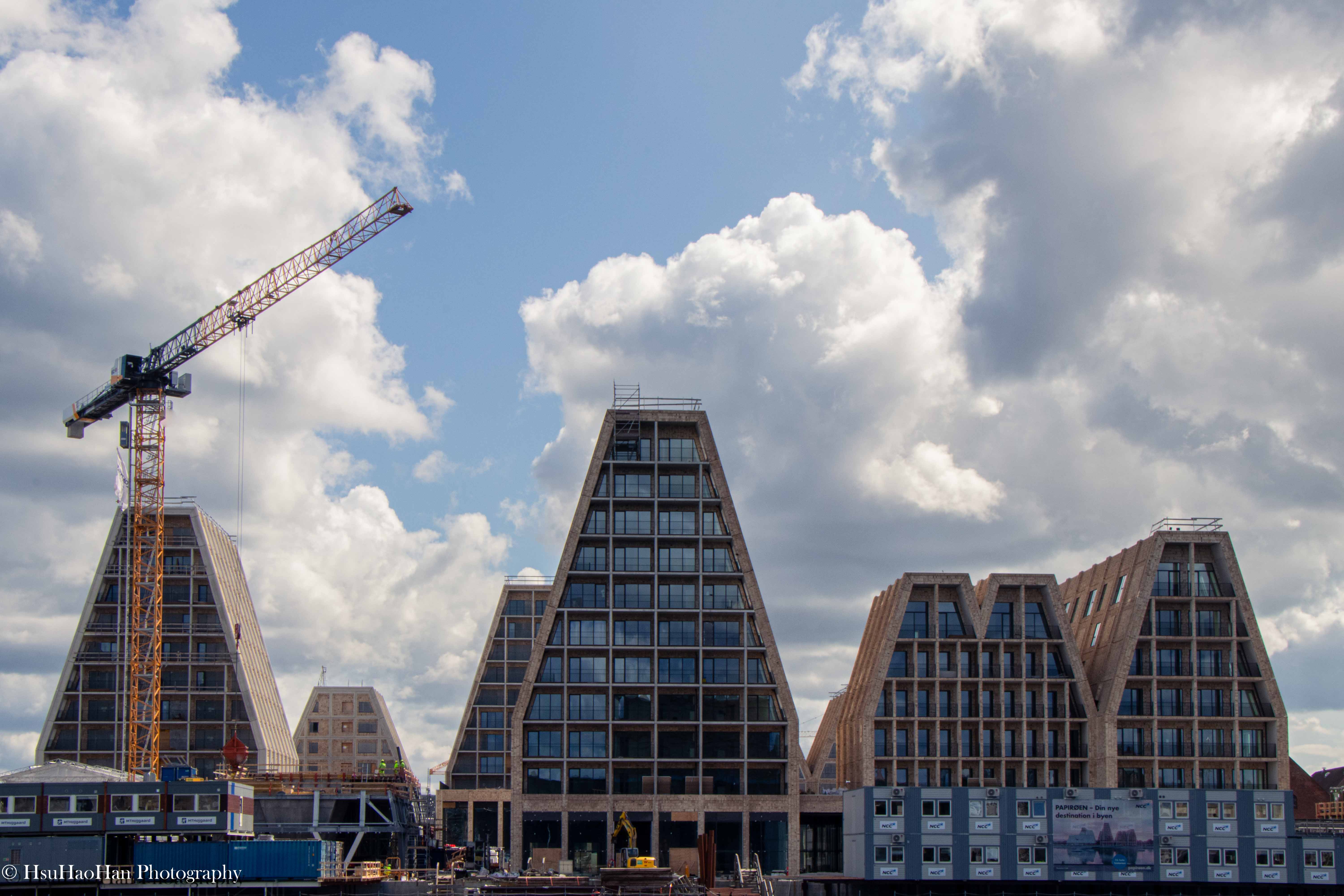 丹麥哥本哈根「紙島」新建案的建築攝影，由COBE設計，數座金字塔形木構造建築在多雲的天空下，前方有起重機正在施工。