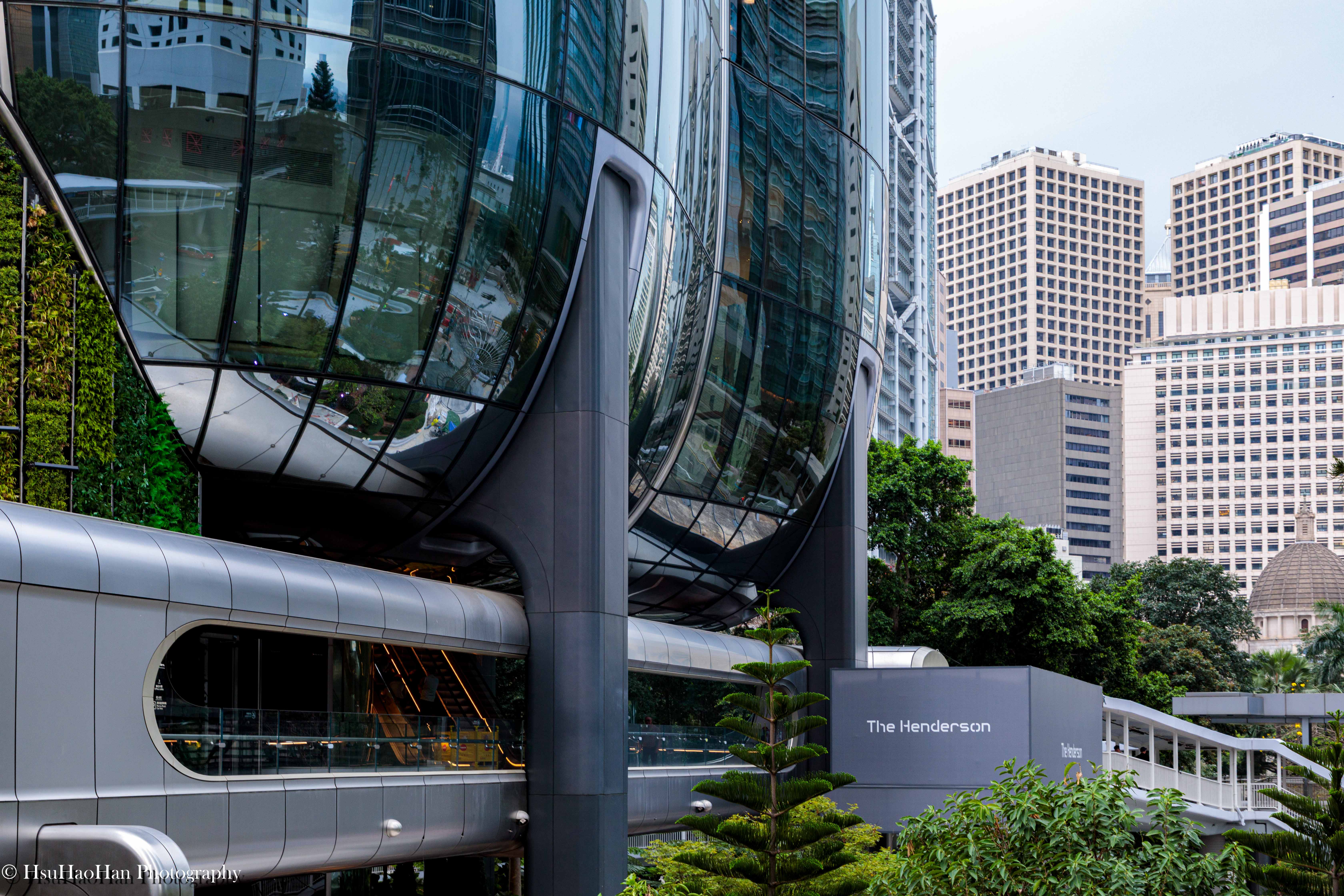 香港 The Henderson 建築的基座細節，巨大的曲面玻璃帷幕反射著周遭的都市景象，下方有天橋與垂直綠化牆面。