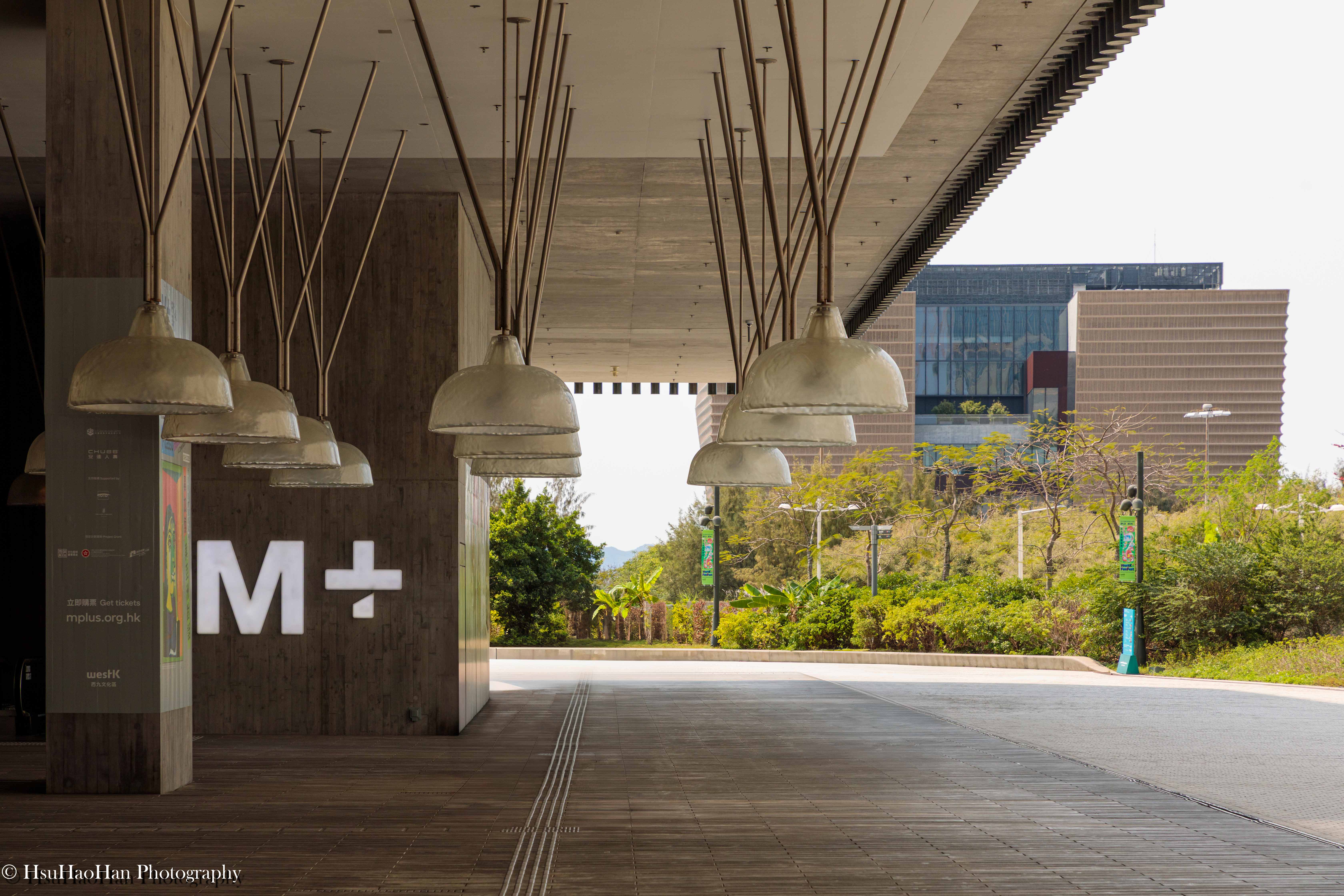 香港 M+ 博物館的入口，在清水混凝土天花板下，懸掛著一排排燈具，M+ 的標誌顯示在木質牆面上。