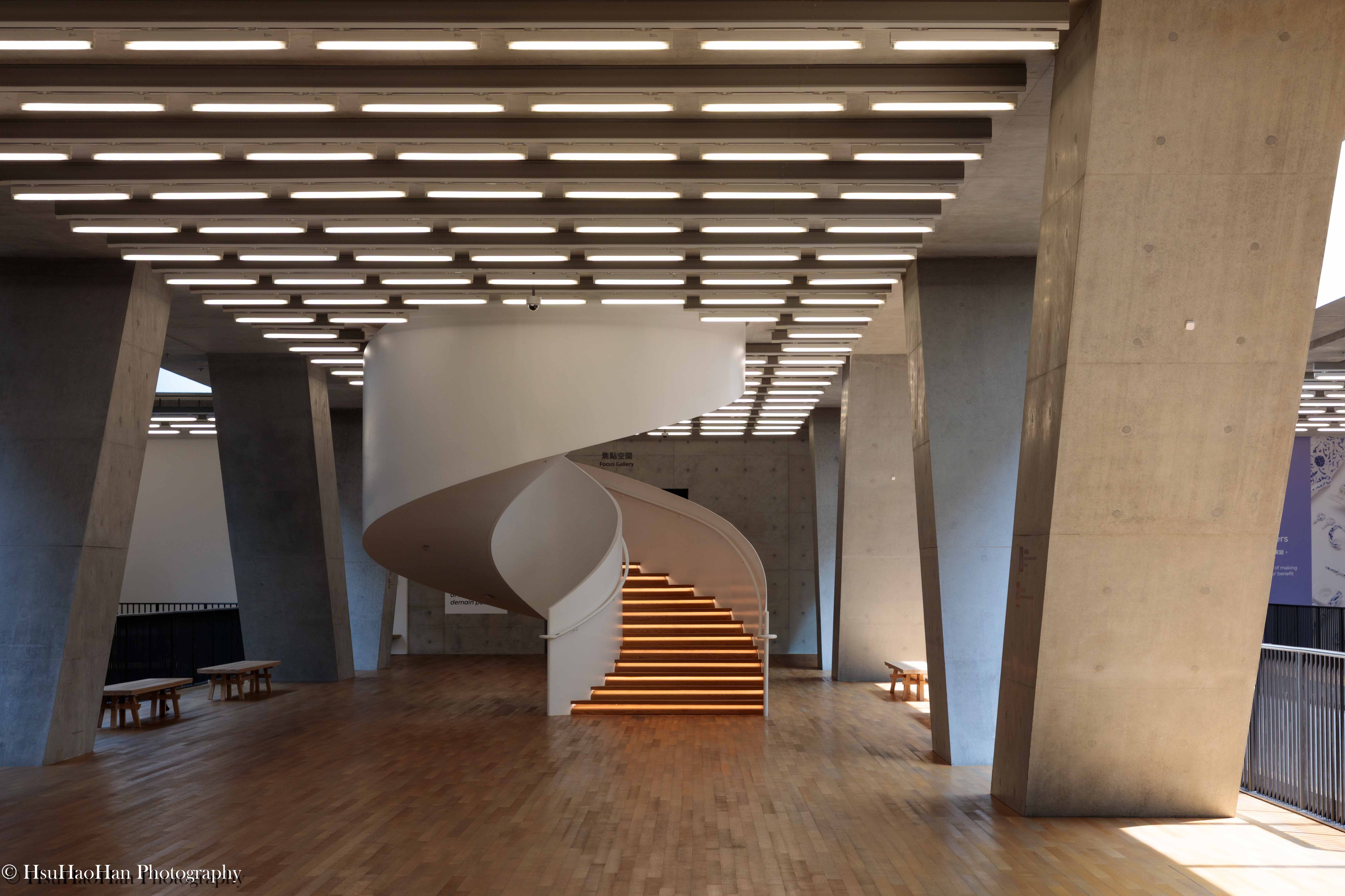 香港 M+ 博物館內的白色螺旋梯，在清水混凝土的巨大支柱之間，樓梯的木質踏階在燈光下發出溫暖的光芒。