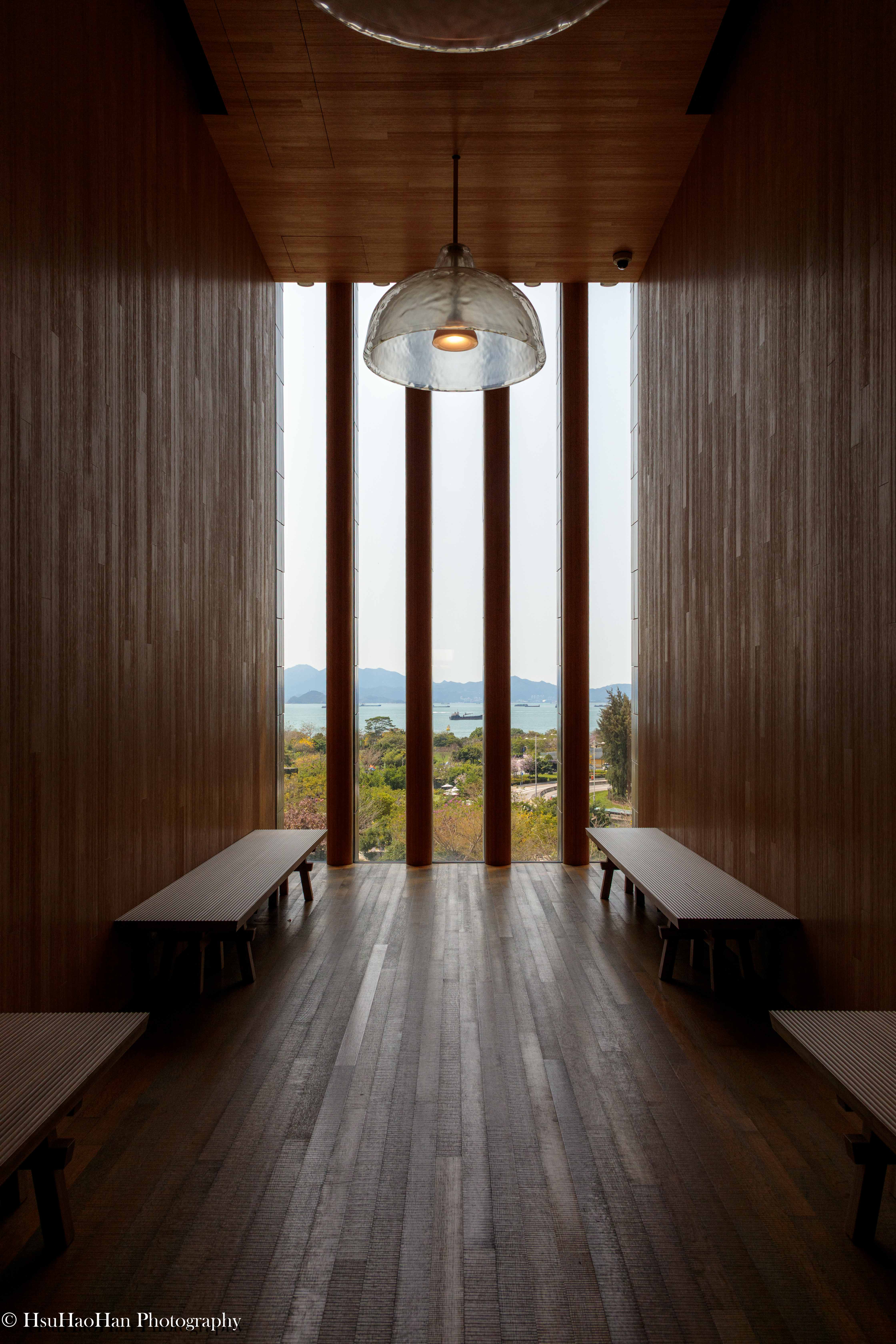 香港 M+ 博物館內的觀景長廊，在對稱的木質牆面之間，透過巨大的落地窗可以看見遠方的海景與島嶼。