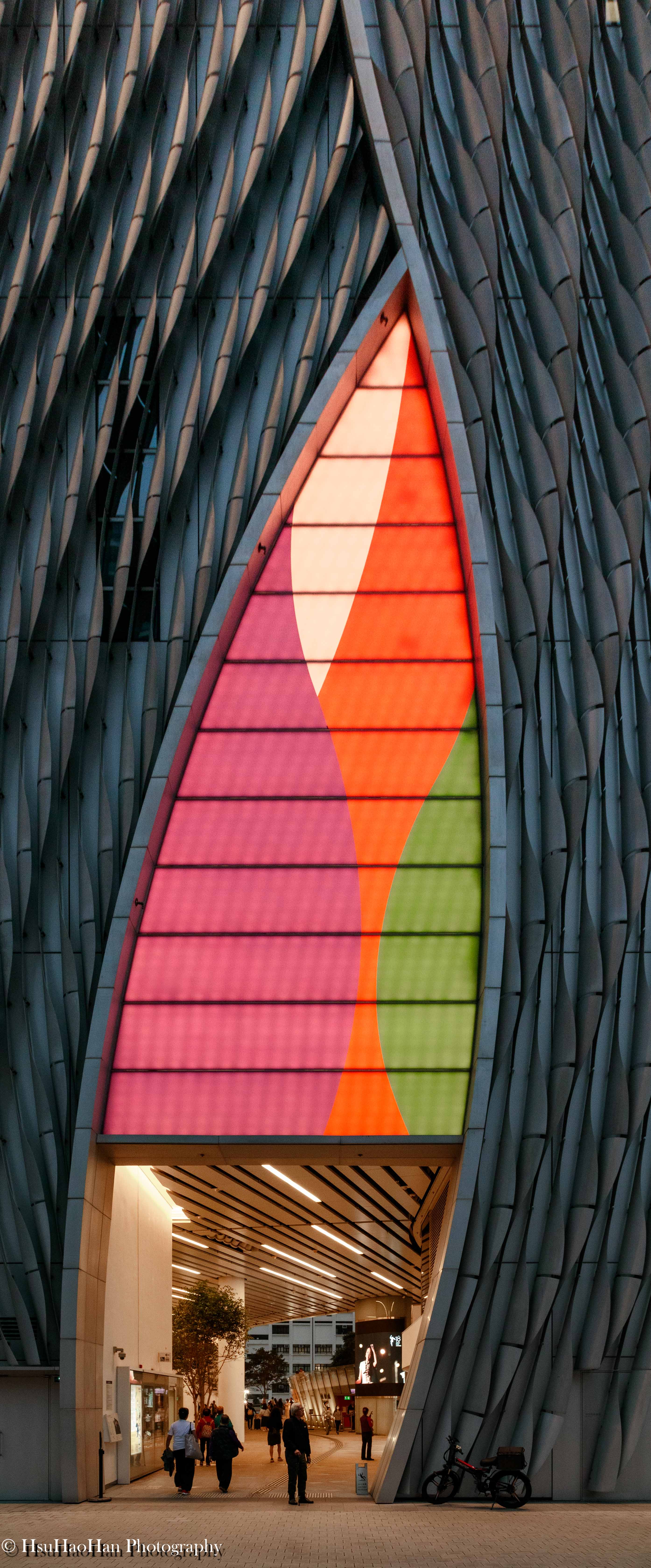 香港戲曲中心的入口特寫，金屬帷幕在底部形成一個巨大的開口，內部透出彩色的燈光與人來人往的通道。