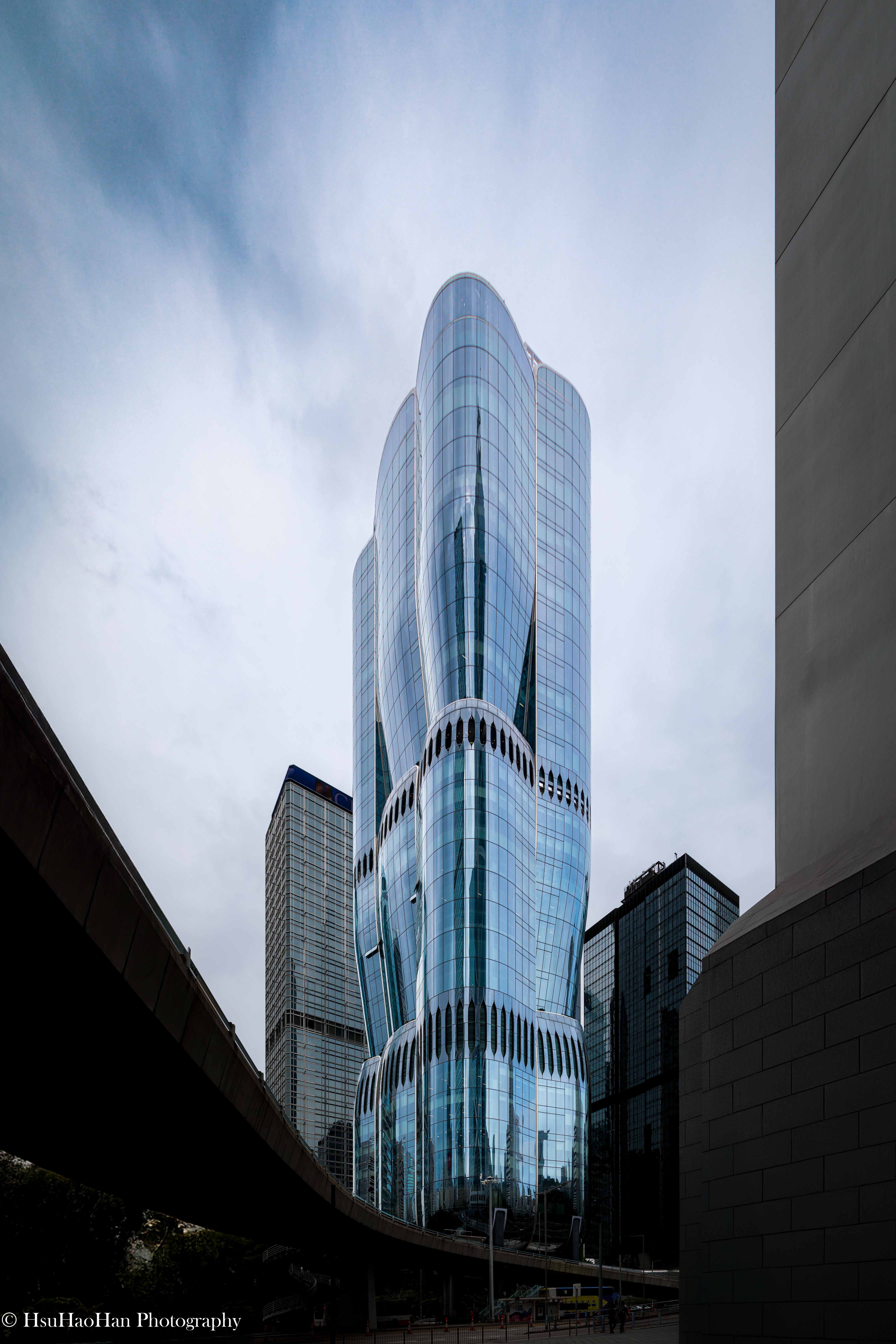 從低角度透過都市天橋框景，仰望香港 The Henderson 大樓，其如花瓣般的曲面玻璃帷幕向上延伸至天空。