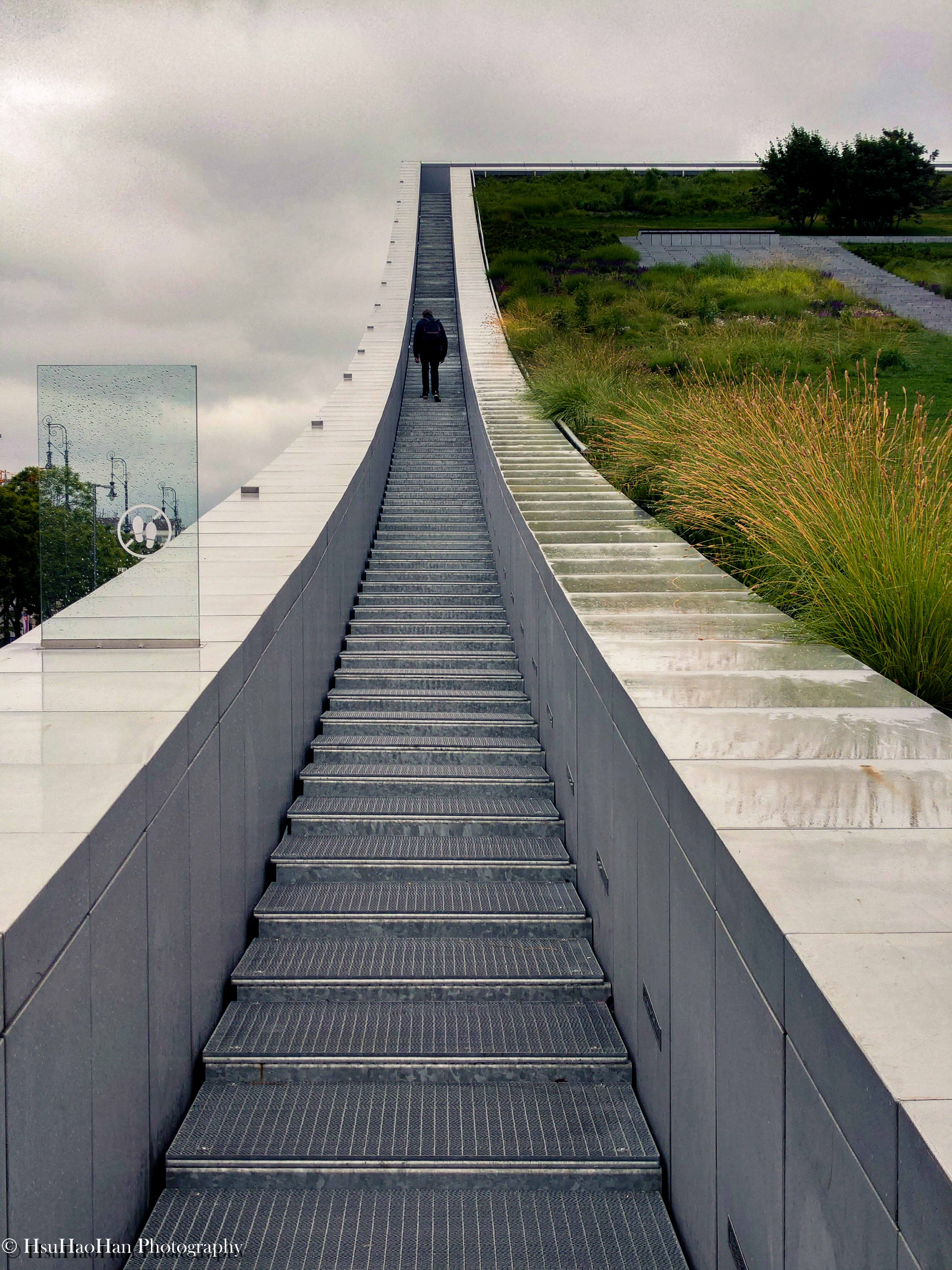 匈牙利布達佩斯民族學博物館內的現代建築攝影，一條灰色金屬階梯向上延伸，一位行人正獨自攀登，周圍是簡約的建築線條和戶外植被。