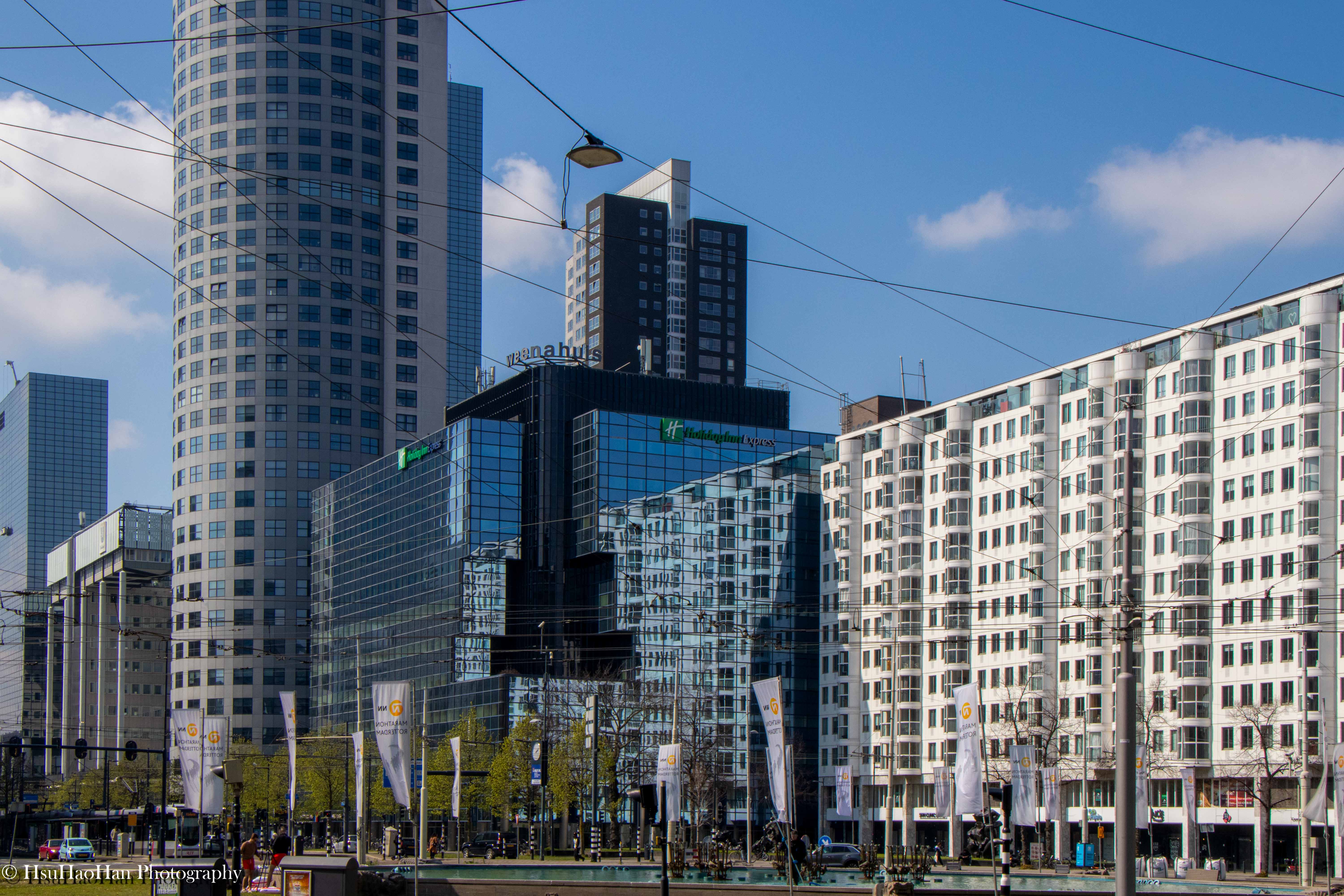 荷蘭鹿特丹市中心的現代街景，畫面中有玻璃帷幕大樓、高聳的住宅塔樓，以及交錯的電車纜線。