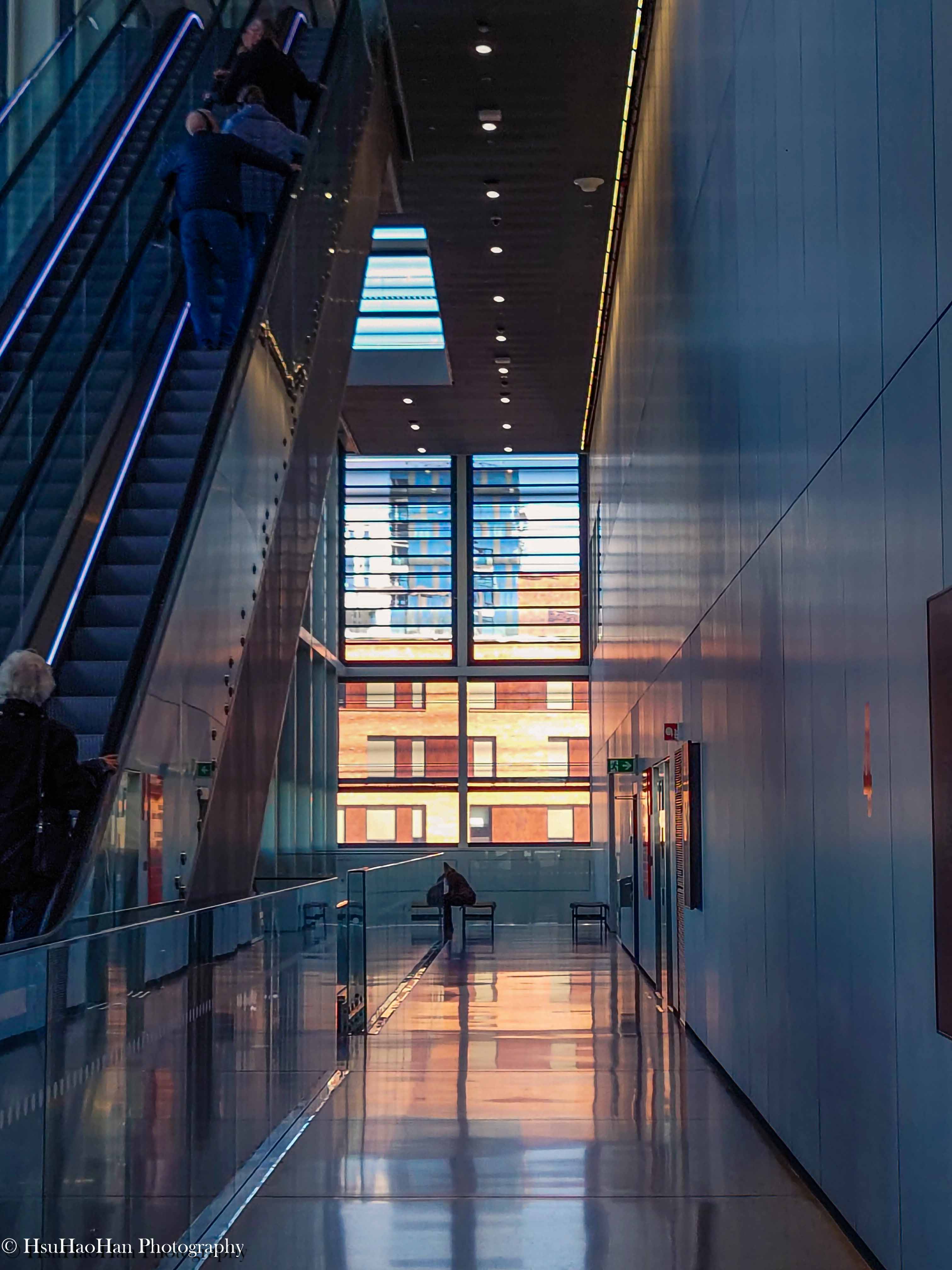 挪威奧斯陸孟克博物館的室內建築攝影，在現代風格的灰色長廊中，訪客正搭乘手扶梯向上，遠處窗邊坐著一個身影，夕陽在地板上投下金色光輝。