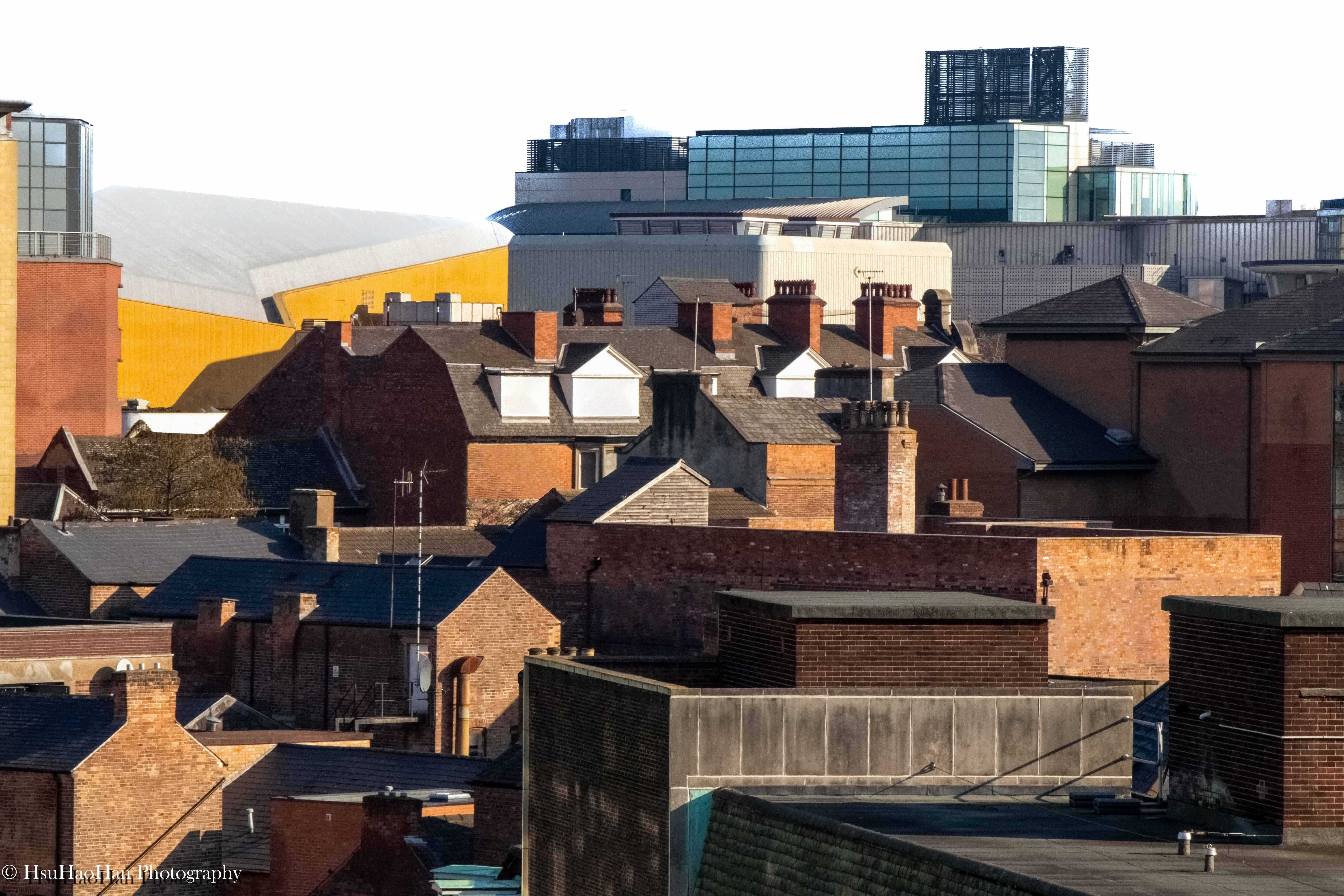 徐浩瀚「城市再現」系列:英國諾丁漢的城市天際線,前景為紅磚屋頂與空調設備,遠景可見不同材質與色彩的新舊建築群。