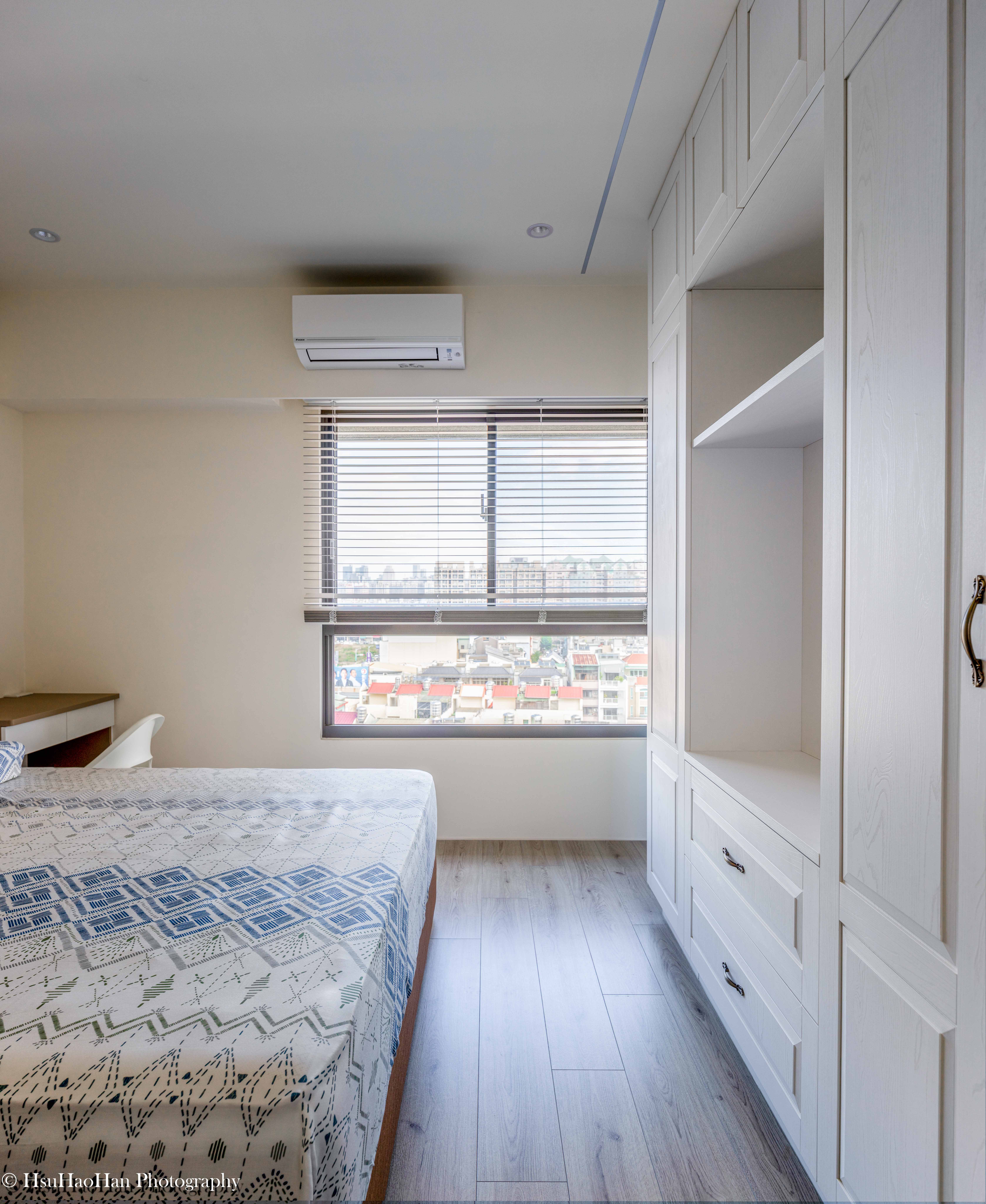 台中「黎民觀邸」的主臥室，床鋪靠著大面窗，窗外是城市景觀，右側是整面的白色系統衣櫃，空間簡潔明亮。