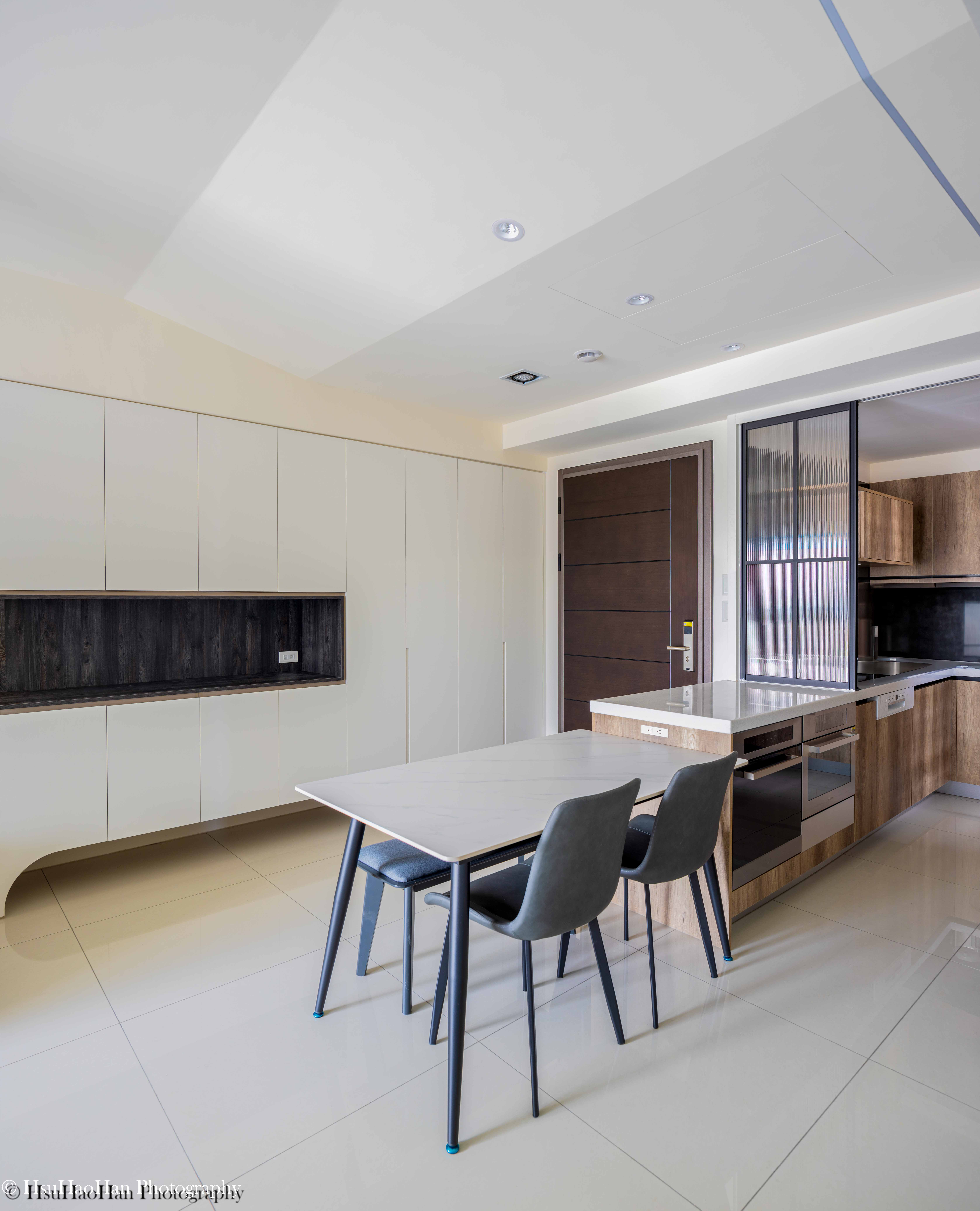 台中「黎民觀邸」的餐廳與廚房中島，白色中島與淺色木紋餐桌相連，搭配灰色餐椅，背景是整面的白色收納櫃。