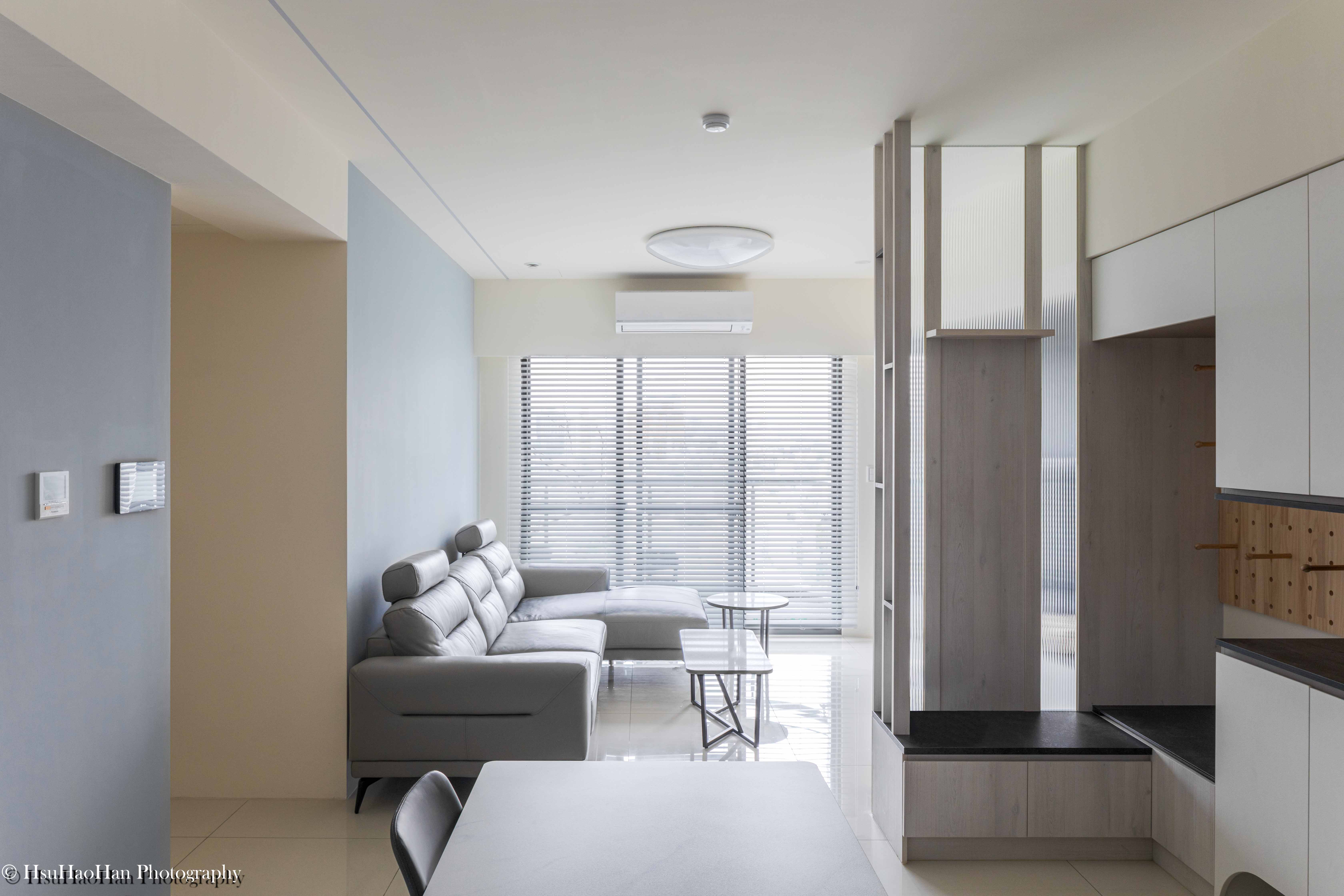 台中「黎民觀邸」的客廳全景，淺灰色L型沙發面向大面落地窗，陽光透過百葉窗灑入，右側是分隔玄關的淺木色隔屏。