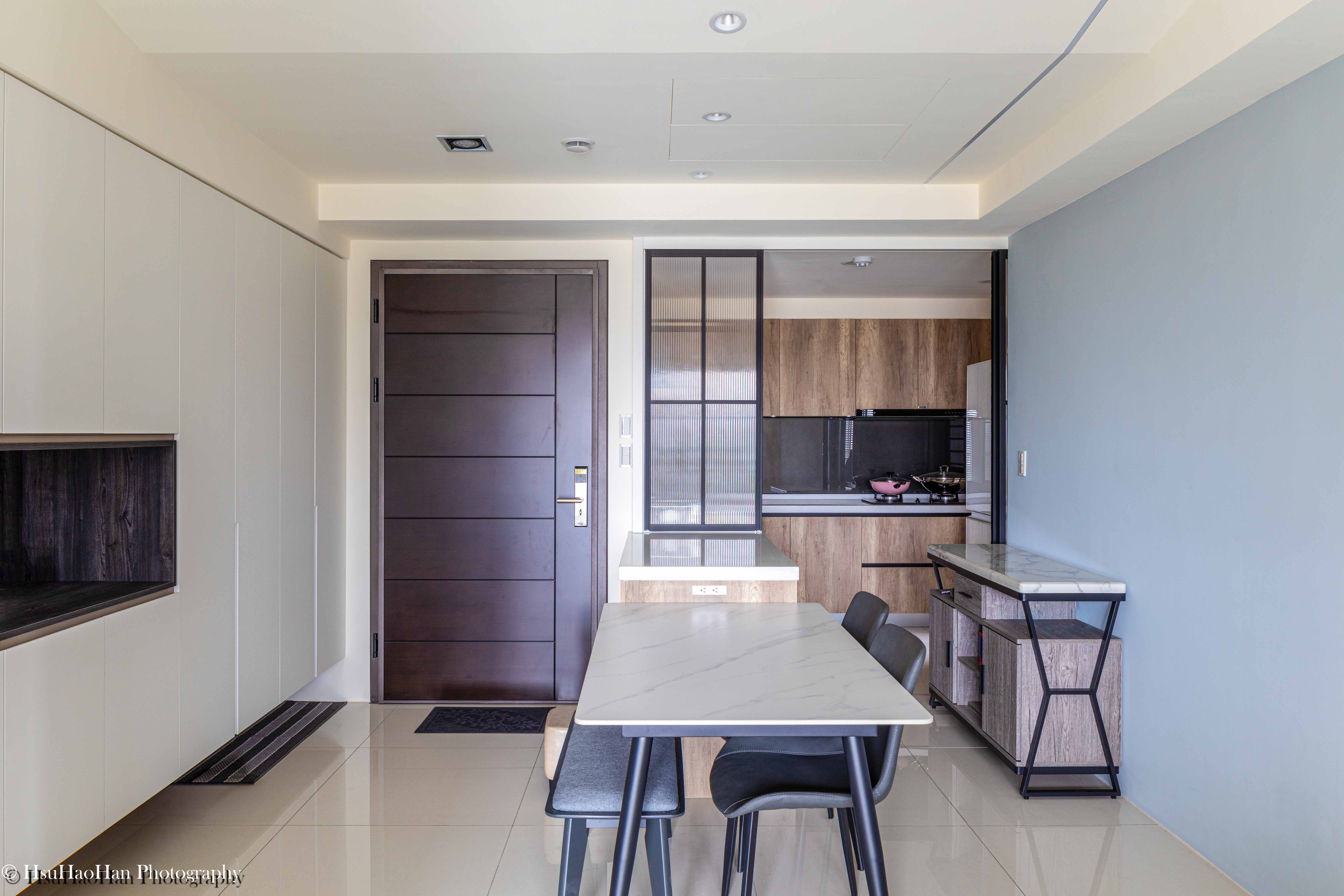 台中「黎民觀邸」的餐廳與廚房中島，白色中島與淺色木紋餐桌相連，搭配灰色餐椅，背景是整面的白色收納櫃。