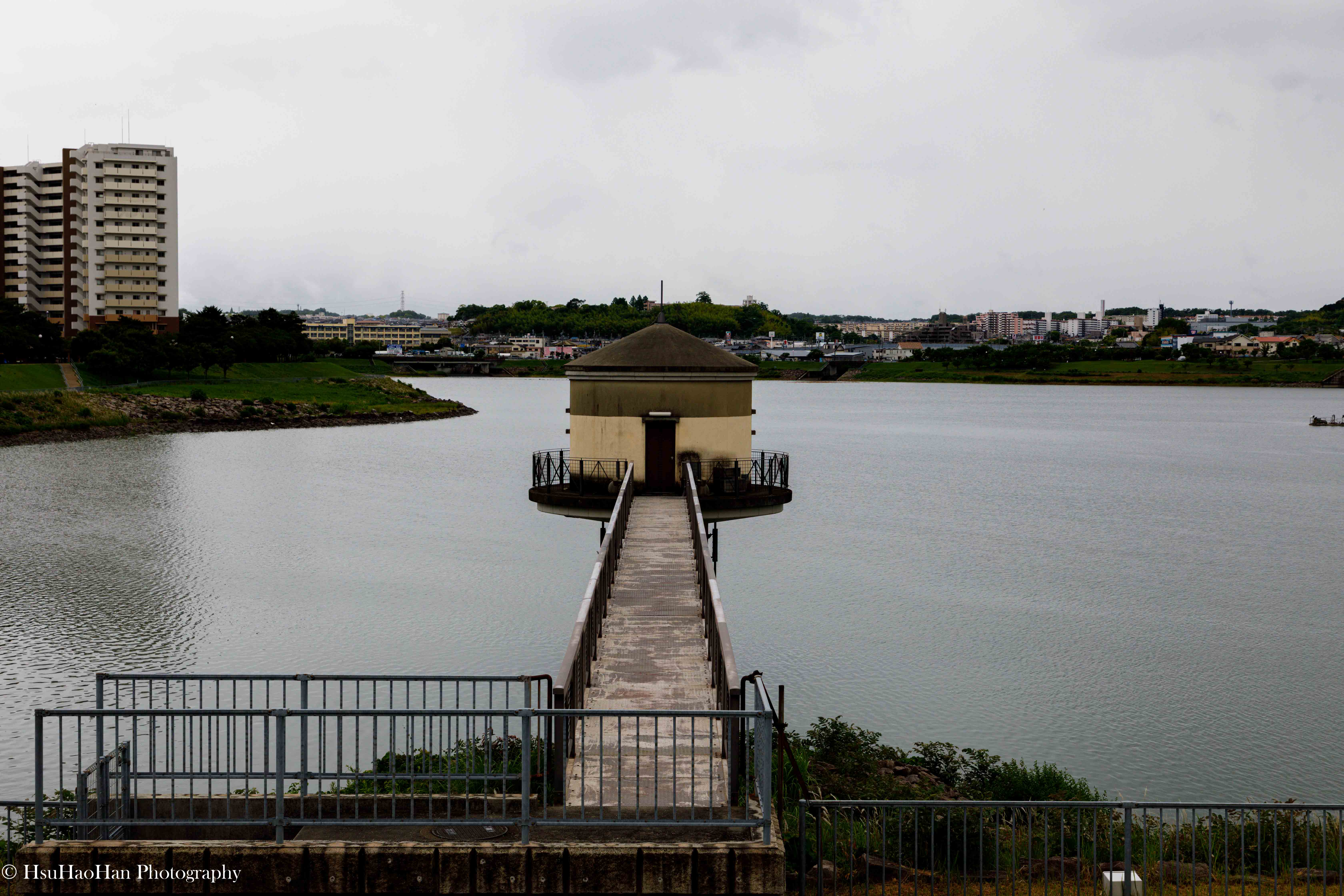 日本大阪狹山池公園的景象，一座長長的棧道延伸至水庫中央的小型圓頂建築物，背景是城市建築與陰天。