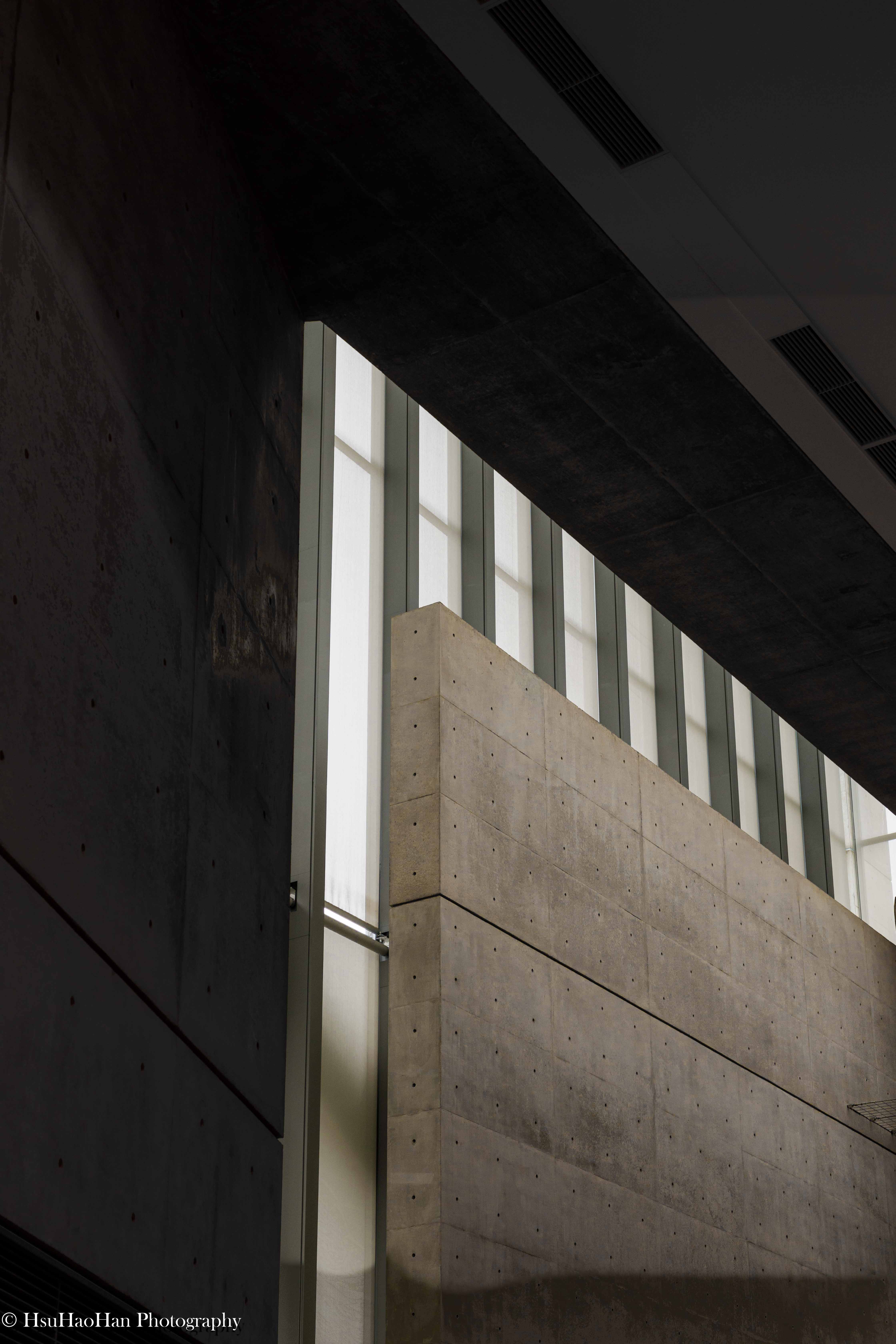 狹山池博物館內部，光線從高處的狹長開口灑落，在巨大的清水混凝土牆面上形成一道明亮的光帶。