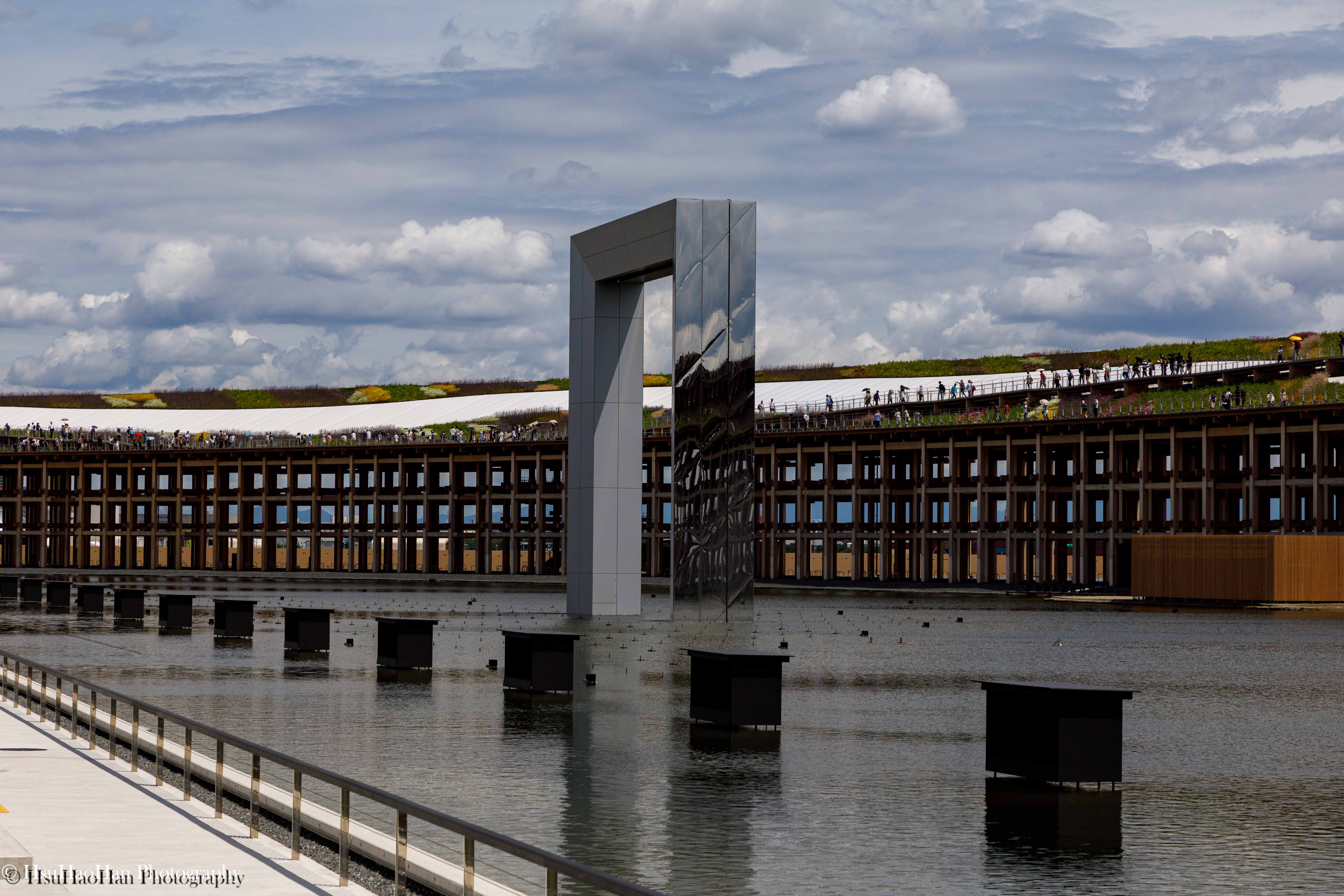 2025大阪萬博會場的水景區域，一個巨大的鏡面拱門裝置矗立在水中，背景是環形屋頂步道與多雲的天空。