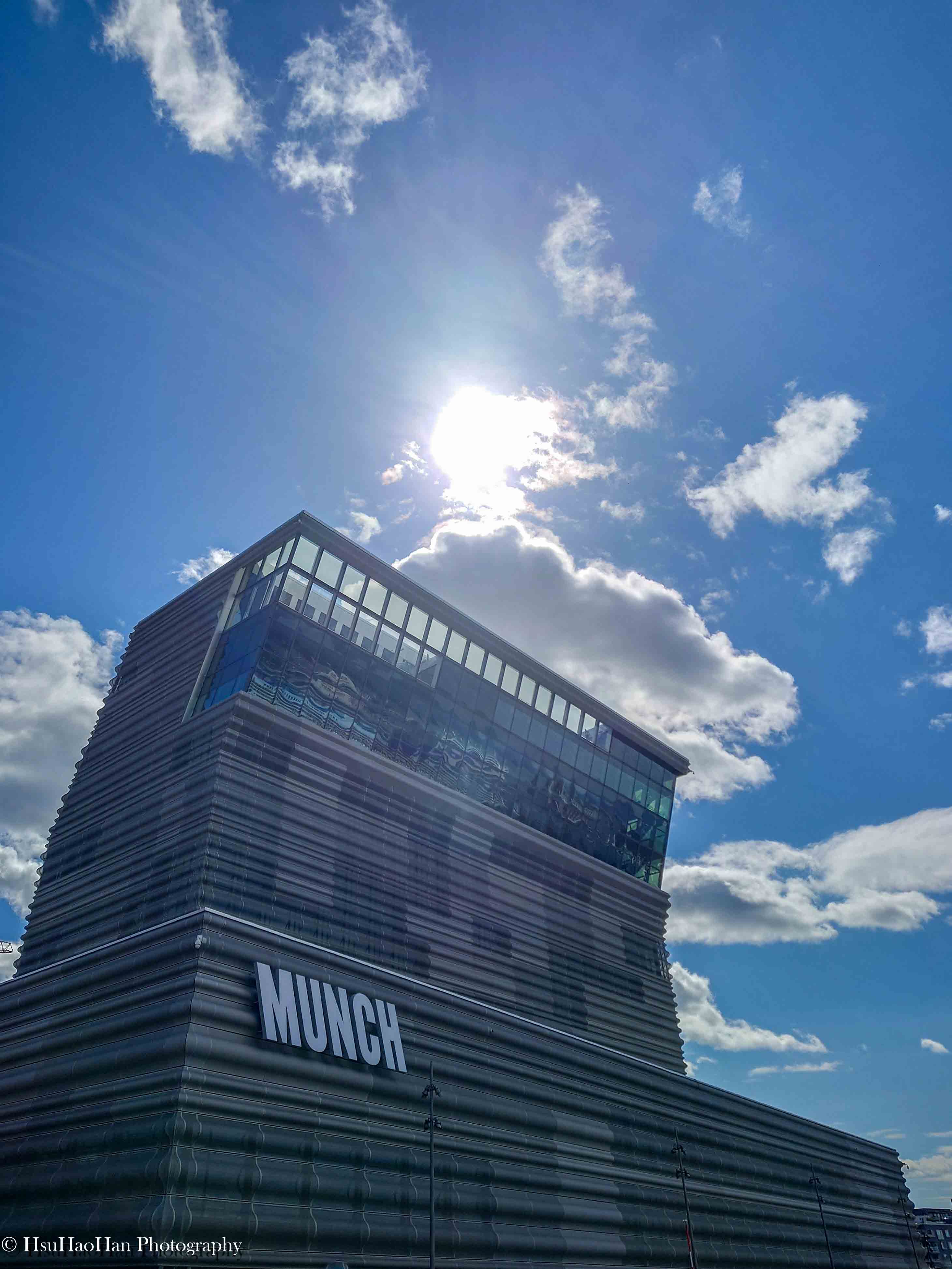 低角度拍攝奧斯陸 Bjørvika 區的 Munch Museum (Munchmuseet)，展示其標誌性的金屬網狀外牆和向外懸挑的結構，陽光穿透雲層照射在頂部。© HsuHaoHan Photography